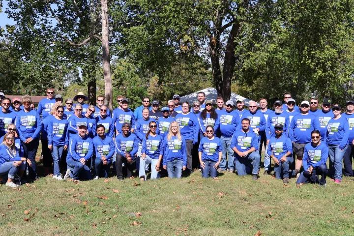 Large group of people wearing matching blue shirts with Day in the Parks printed on them, posing together outdoors in a park on a sunny day, with trees and event tents in the background.