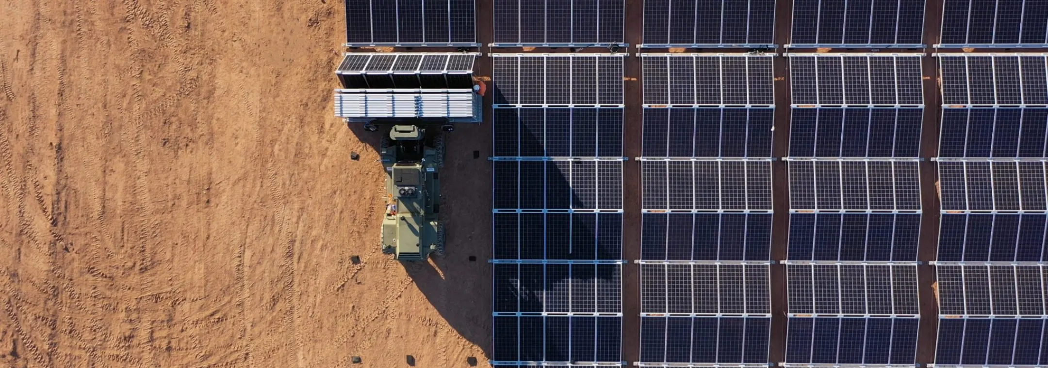 Aerial view of a solar panel installation in a desert area with a vehicle transporting additional panels.