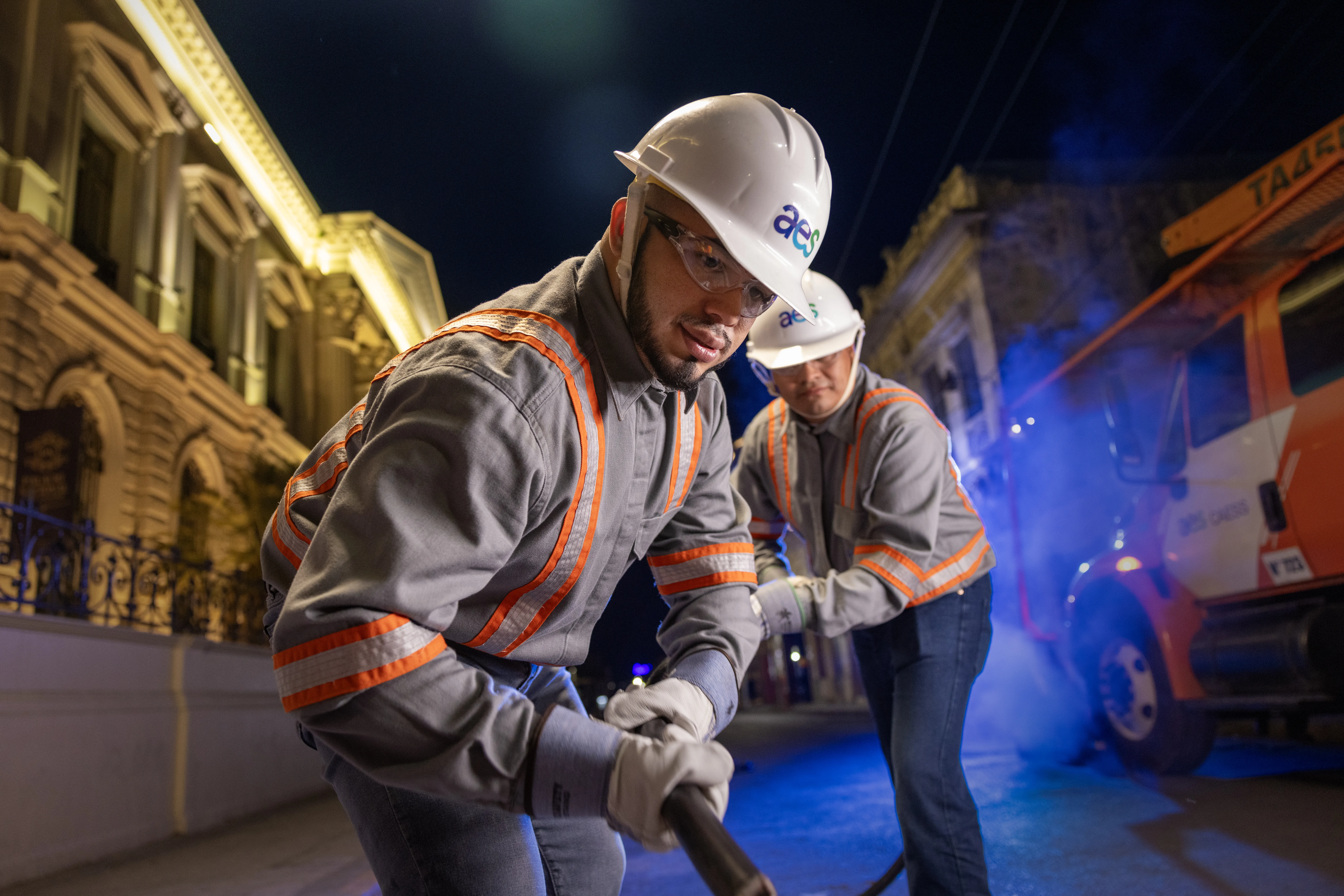 Dos técnicos de AES con equipo de protección completo trabajan durante la noche en una calle urbana. En primer plano, un técnico jala un cable grueso mientras un compañero lo asiste al fondo cerca de un camión de servicio naranja. La escena está iluminada artificialmente frente a un edificio de arquitectura clásica.