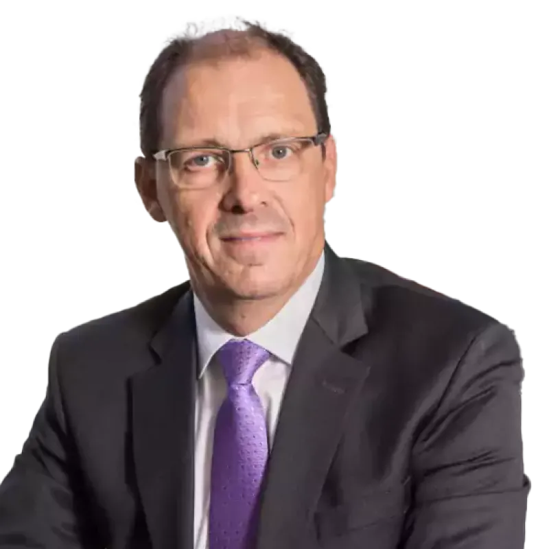 Man wearing glasses and a dark suit with a purple tie, seated in an office with large windows overlooking a cityscape.
