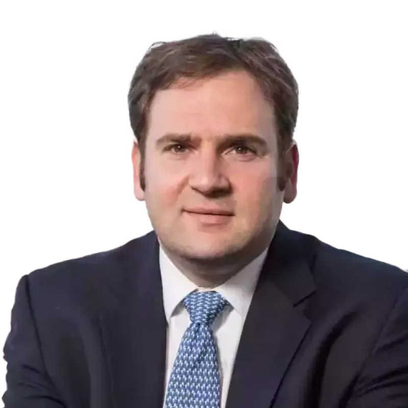 Man in a business suit with blue patterned tie sitting in front of a window with a cityscape in the background.