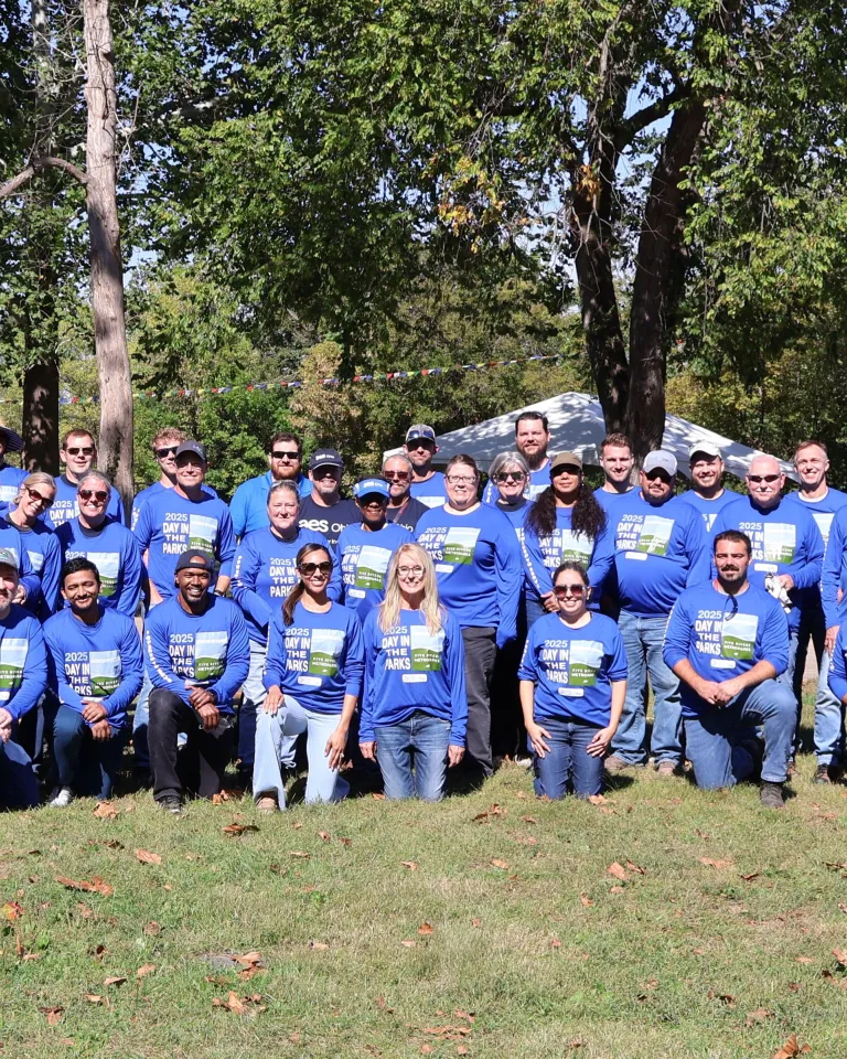Large group of people wearing matching blue shirts with Day in the Parks printed on them, posing together outdoors in a park on a sunny day, with trees and event tents in the background.